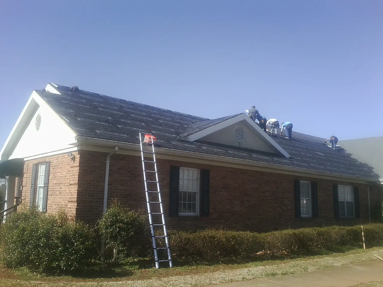 Crew tearing shingles off a brick ranch roof with a ladder set — Emergency Roof Repair work by our crew in Colchester